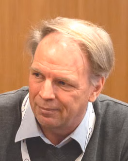 A middle-aged person with light hair wears a grey jumper over a light blue collared shirt and a lanyard with partially visible text. They are standing against a wooden-panelled background, suggesting an indoor setting such as a conference, seminar, or professional gathering. The individual appears engaged and approachable, contributing to a formal yet friendly atmosphere.