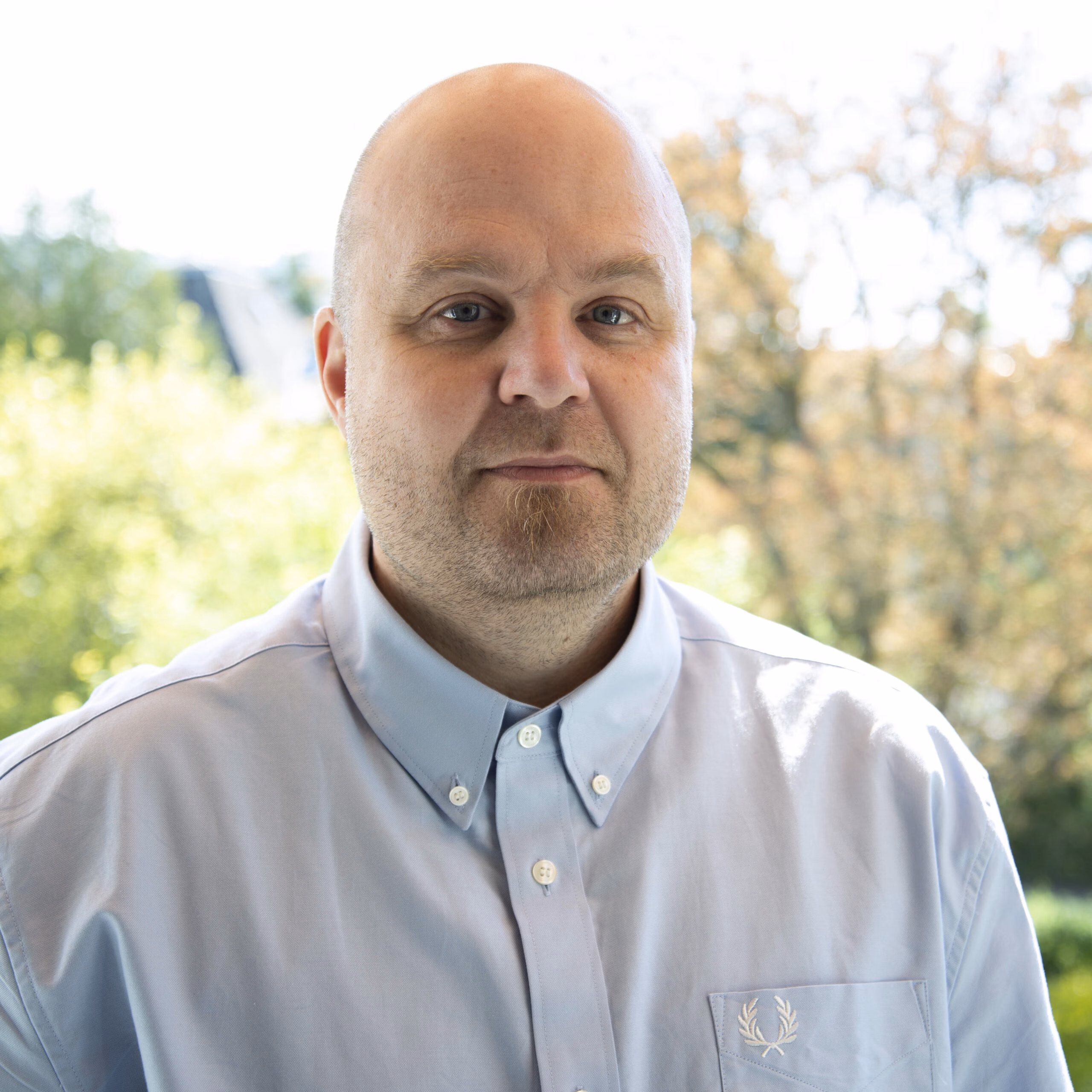 A person with a bald head and neutral expression stands in front of a sunlit window, wearing a light blue button-up shirt with a small Fred Perry laurel wreath logo. Trees and daylight outside create a bright, natural backdrop, adding a calm and composed tone to the portrait.