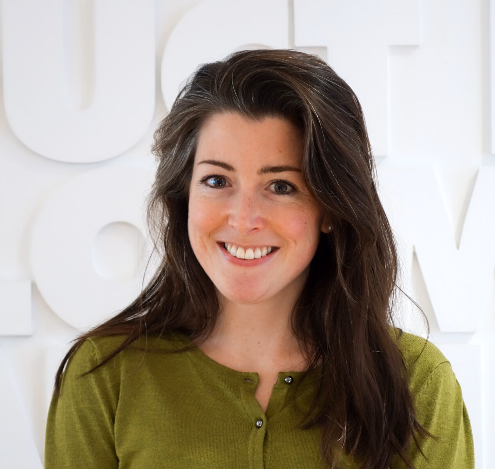 A person with long dark brown hair smiles warmly at the camera. They wear a green buttoned shirt and stand against a white wall featuring large, raised letters that are only partially visible. The setting appears professional yet relaxed, and the individual’s friendly expression adds a welcoming tone to the image.