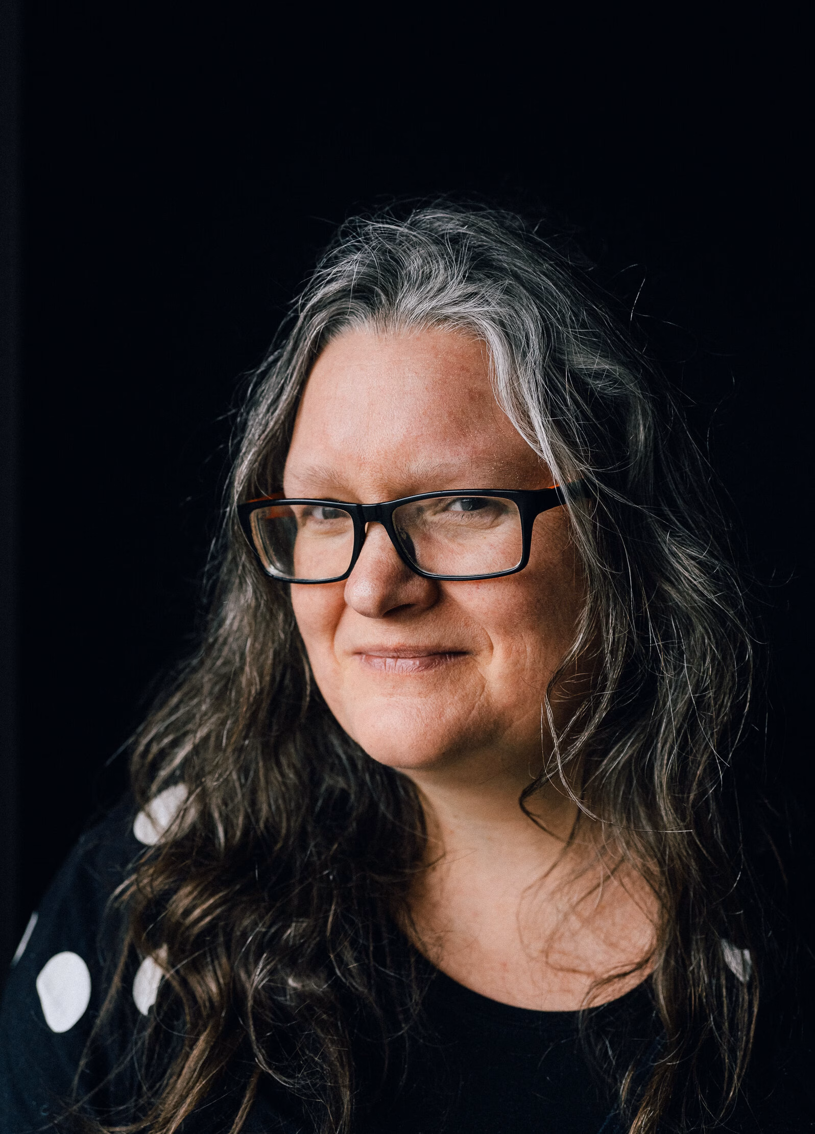 A close-up portrait of a person with long, wavy, greying hair and rectangular glasses, wearing a dark top with white polka dots. The individual is set against a black background, with lighting that accentuates their facial features and expression. The image conveys a natural and thoughtful presence, suggesting authenticity and introspection.