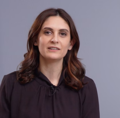 A person with long wavy brown hair wears a dark blouse and looks directly at the camera with a neutral expression. Set against a plain light grey background, the portrait has a calm and composed tone, highlighting the subject’s natural presence.