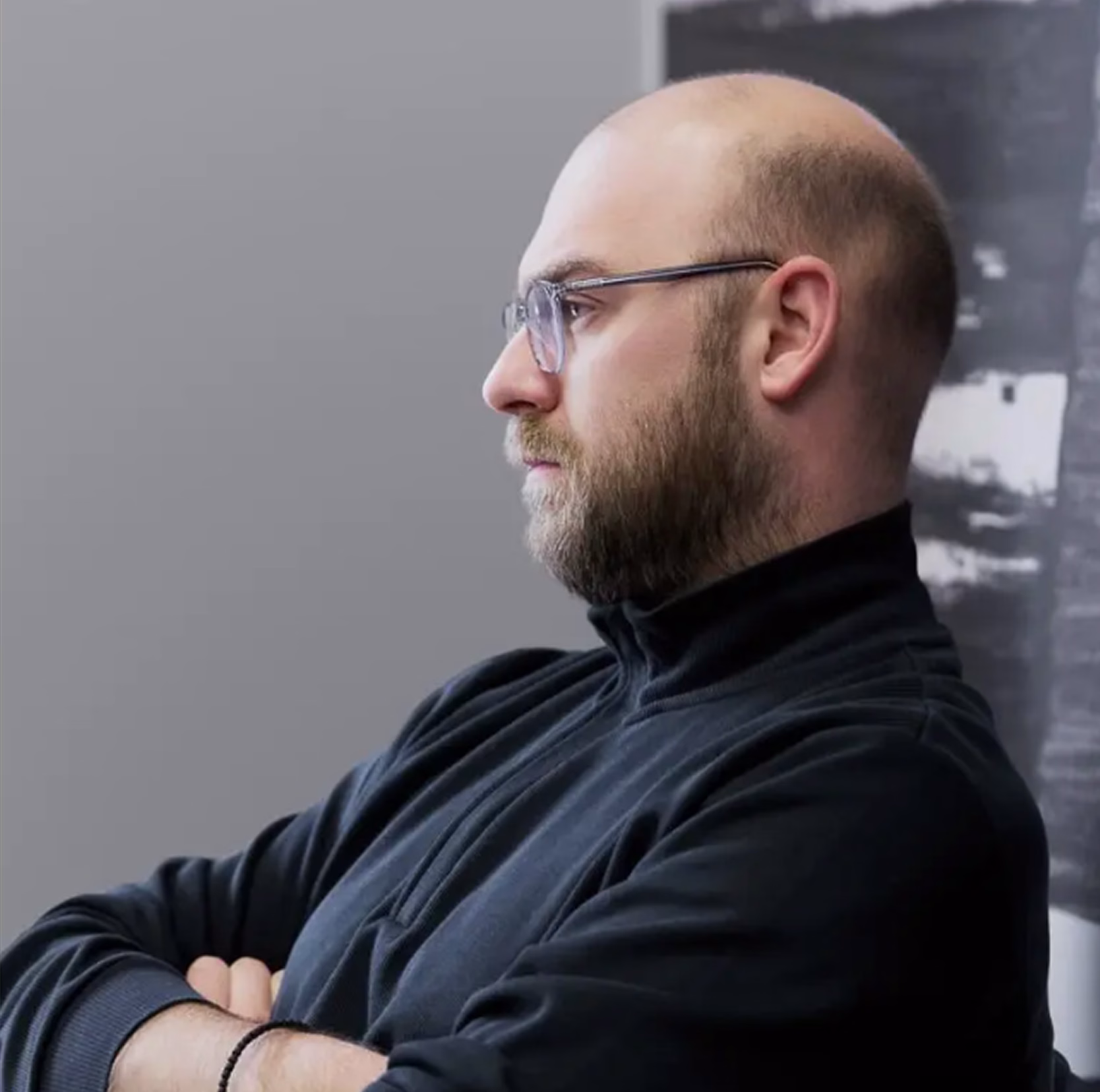 A bald man with a beard, glasses, and a black zip-up jacket sits or stands in profile with arms crossed, gazing thoughtfully to the left. The background features a grey wall and a partially visible black-and-white abstract artwork, creating a calm, introspective atmosphere