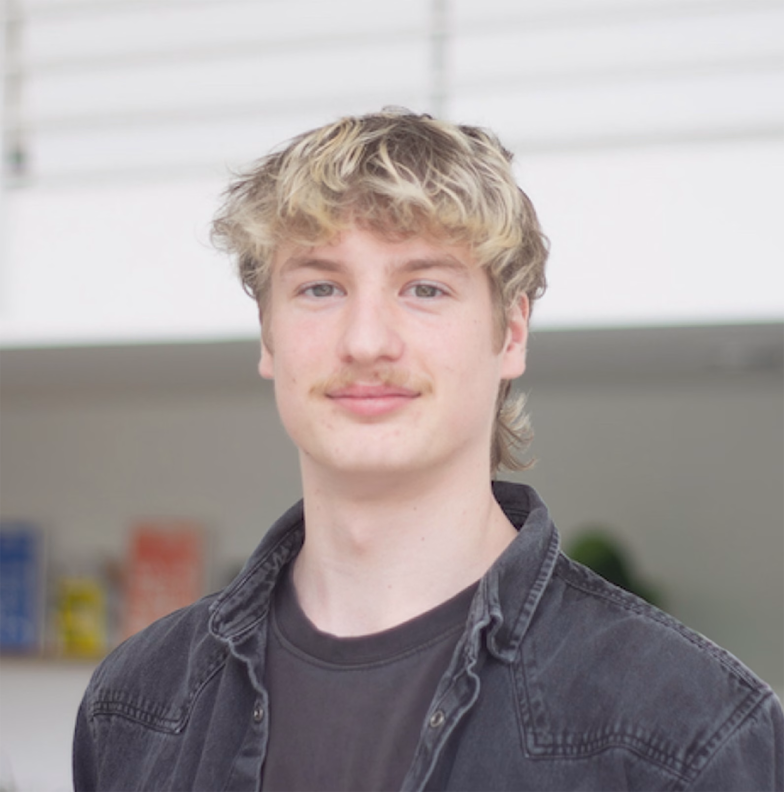 A person with wavy light-coloured hair and a moustache wears a black denim jacket over a black t-shirt, standing indoors near a white railing. The background includes softly blurred shelves with books or containers. Bright natural light gives the scene a relaxed, candid feel.