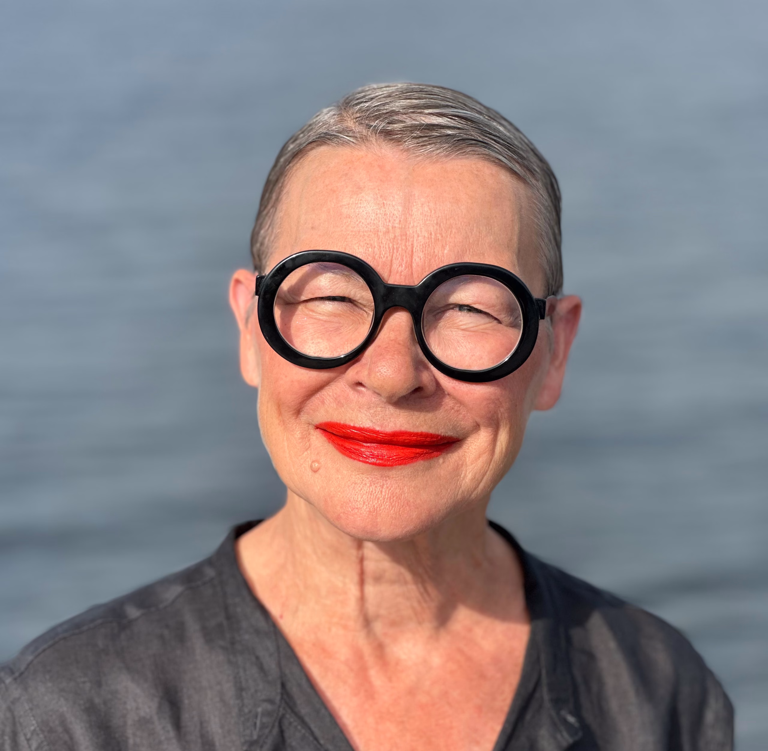 An older person with short, neatly combed grey hair stands outdoors in front of a body of water. They wear large round black glasses, bright red lipstick, and a dark top. Sunlight illuminates their face as they squint slightly, with the serene water backdrop highlighting their bold and expressive style.
